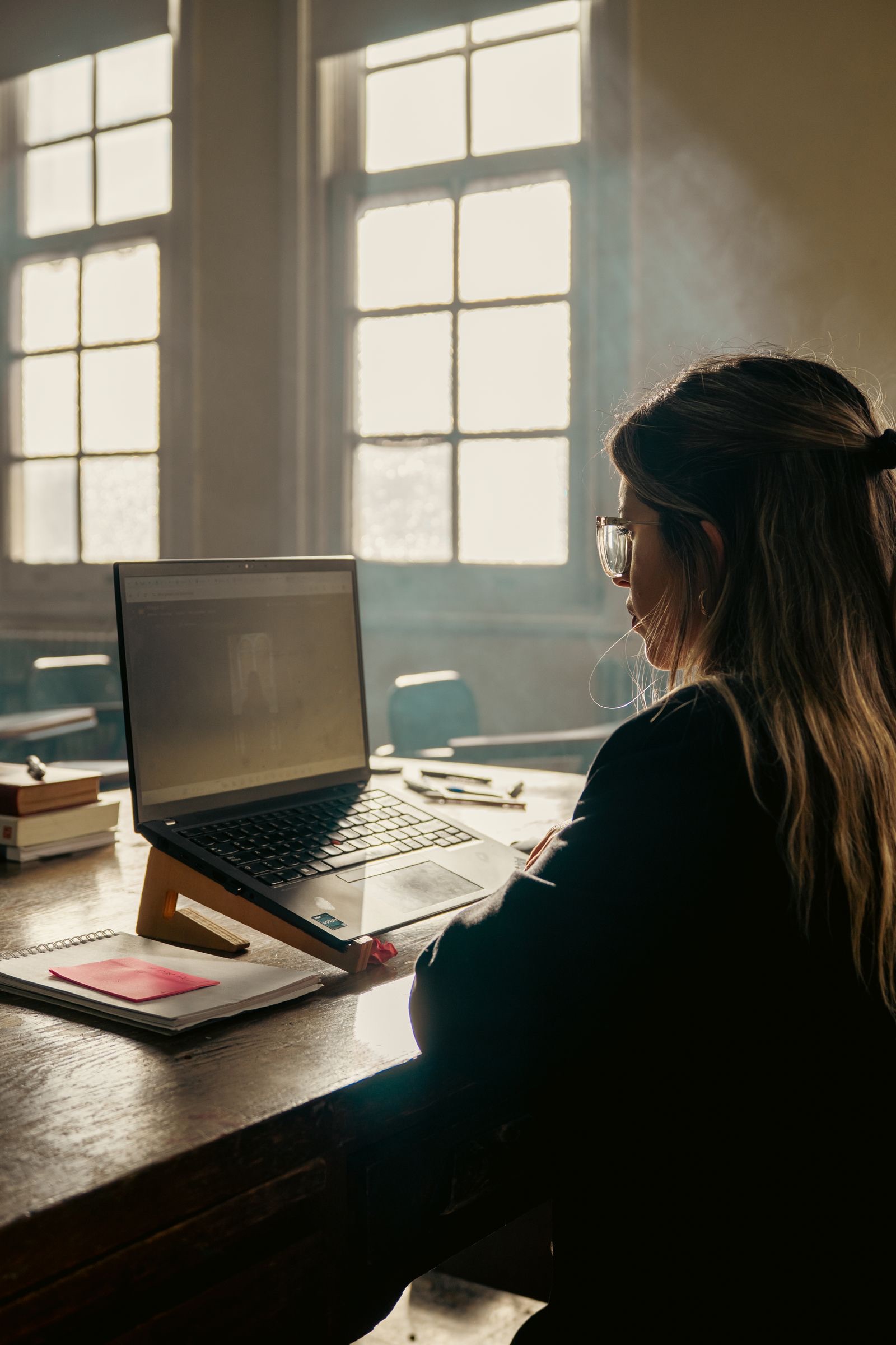 A student works on a laptop at a desk in a sunlit classroom with large windows