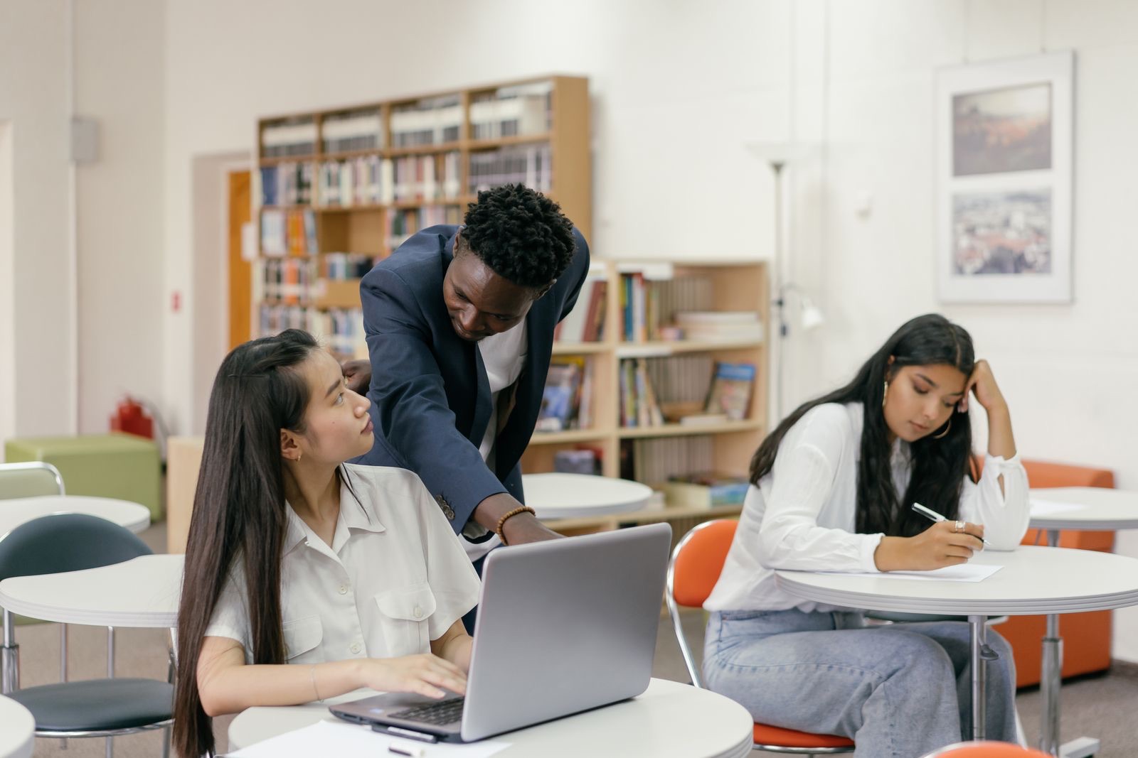 A teacher leans over a student working at a laptop, pointing at the screen